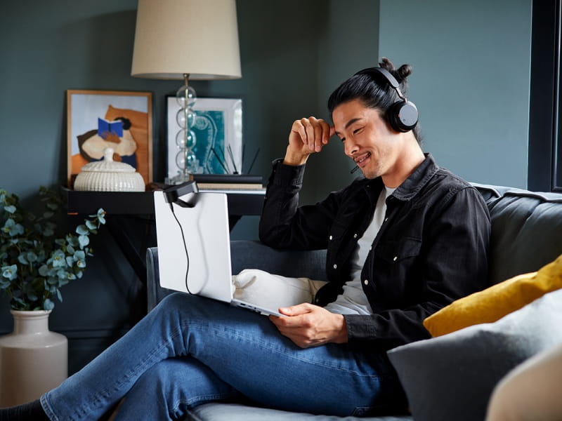 Person wearing headphones, sitting cross-legged on a sofa with a laptop on their lap in a cozy living room.